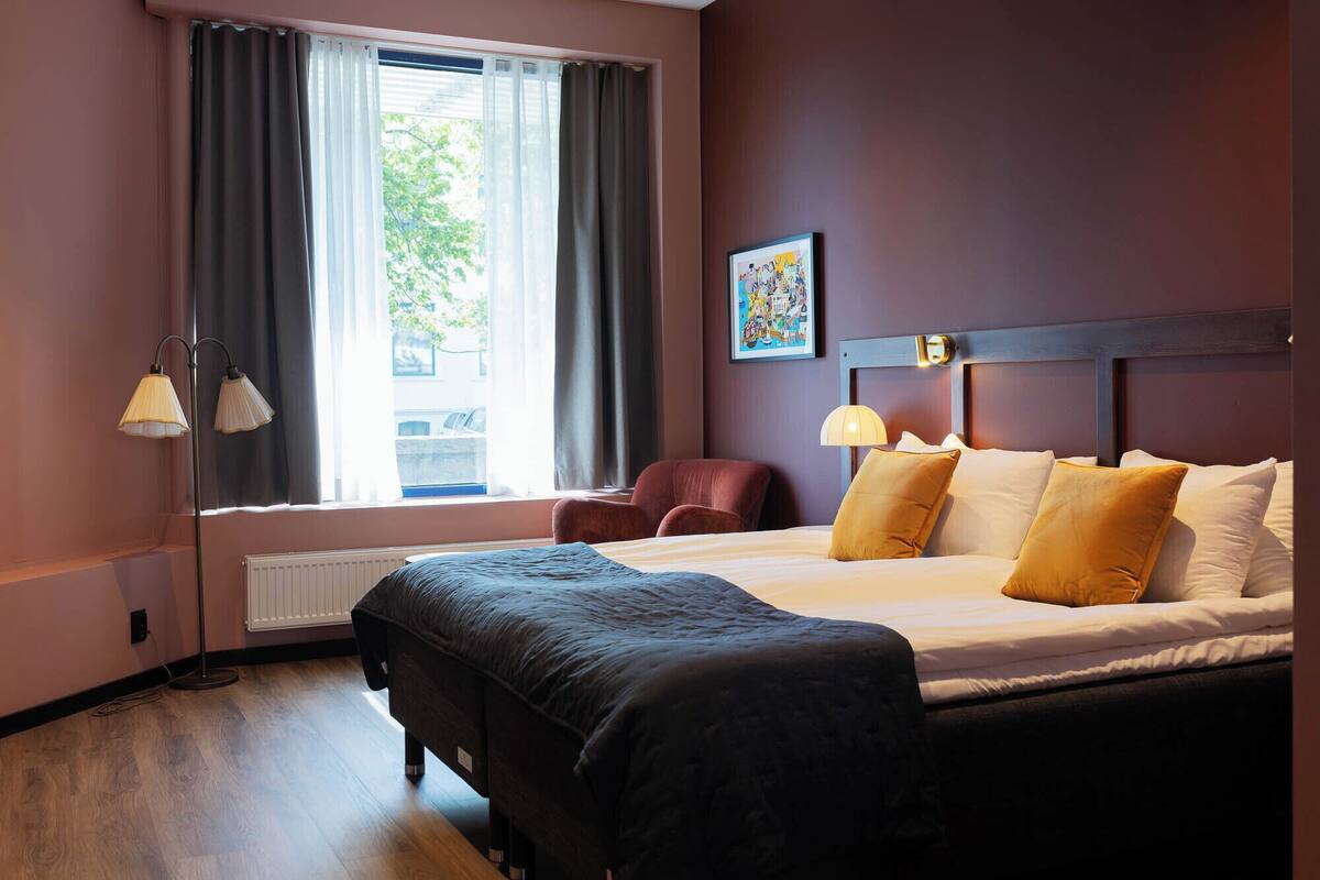 A hotel room with a large bed, two bedside lamps, a chair, a radiator, and a window with curtains.