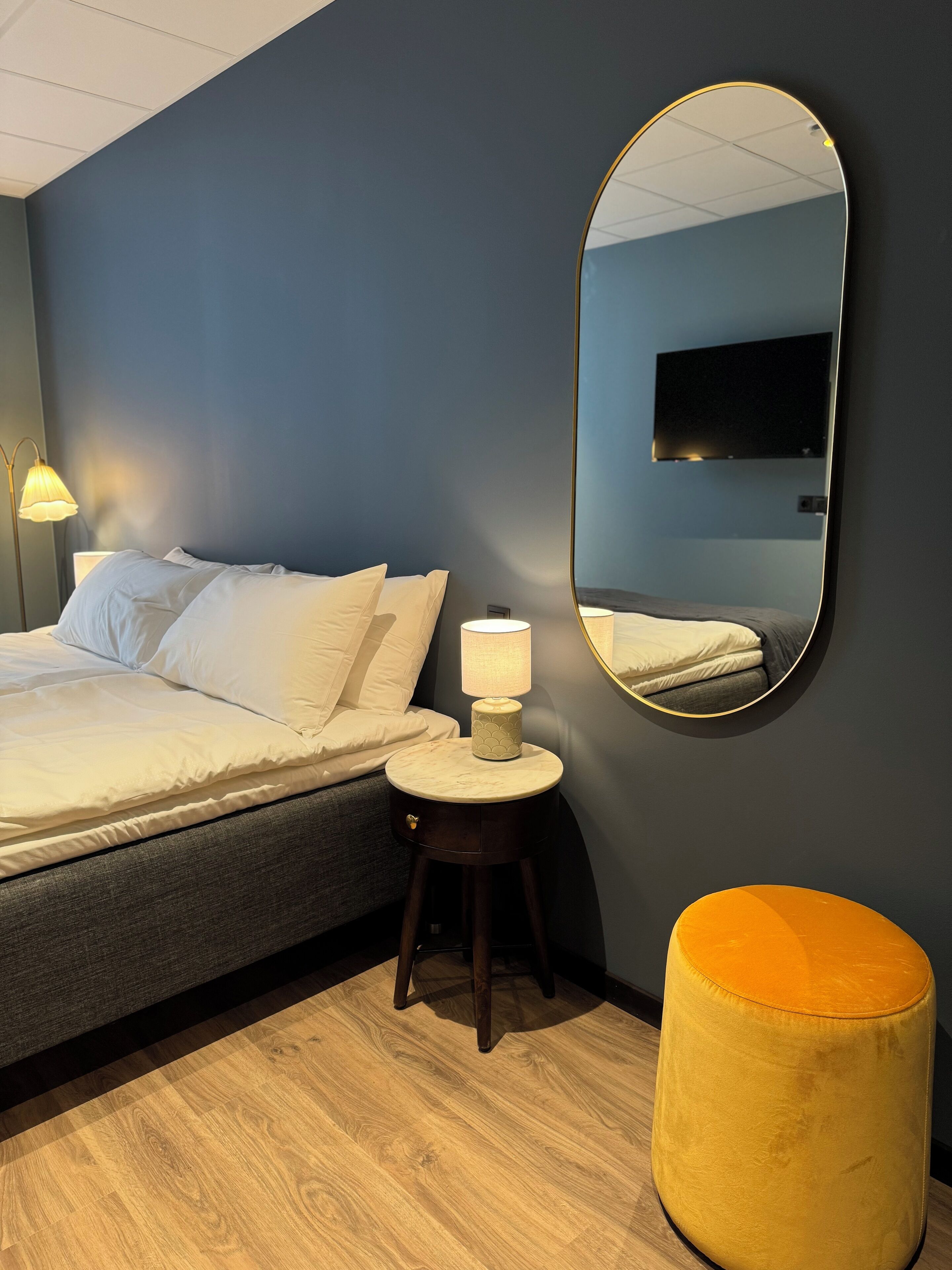 Un dormitorio con cama, lámpara de noche, espejo redondo y suelo de madera.