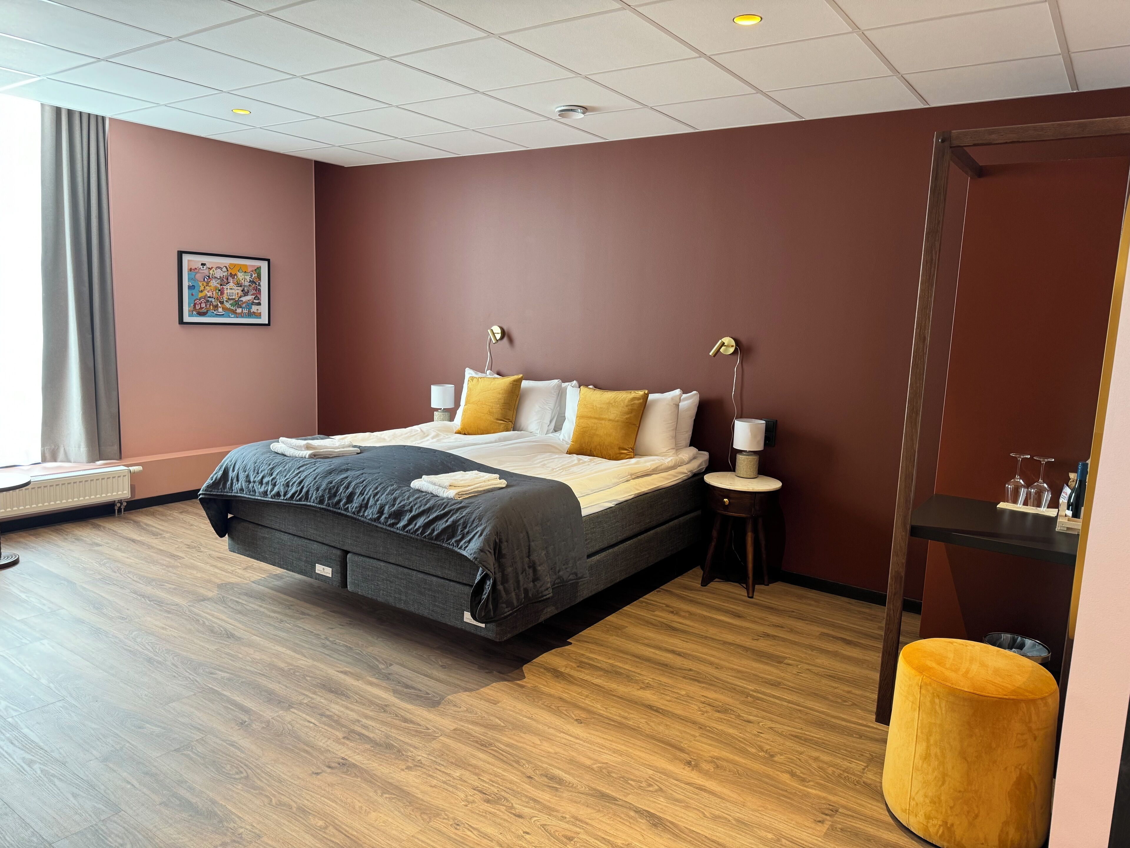Una habitación de hotel moderna con una cama grande, una mesita de noche con lámpara, una pequeña mesa redonda con vasos y un radiador.
