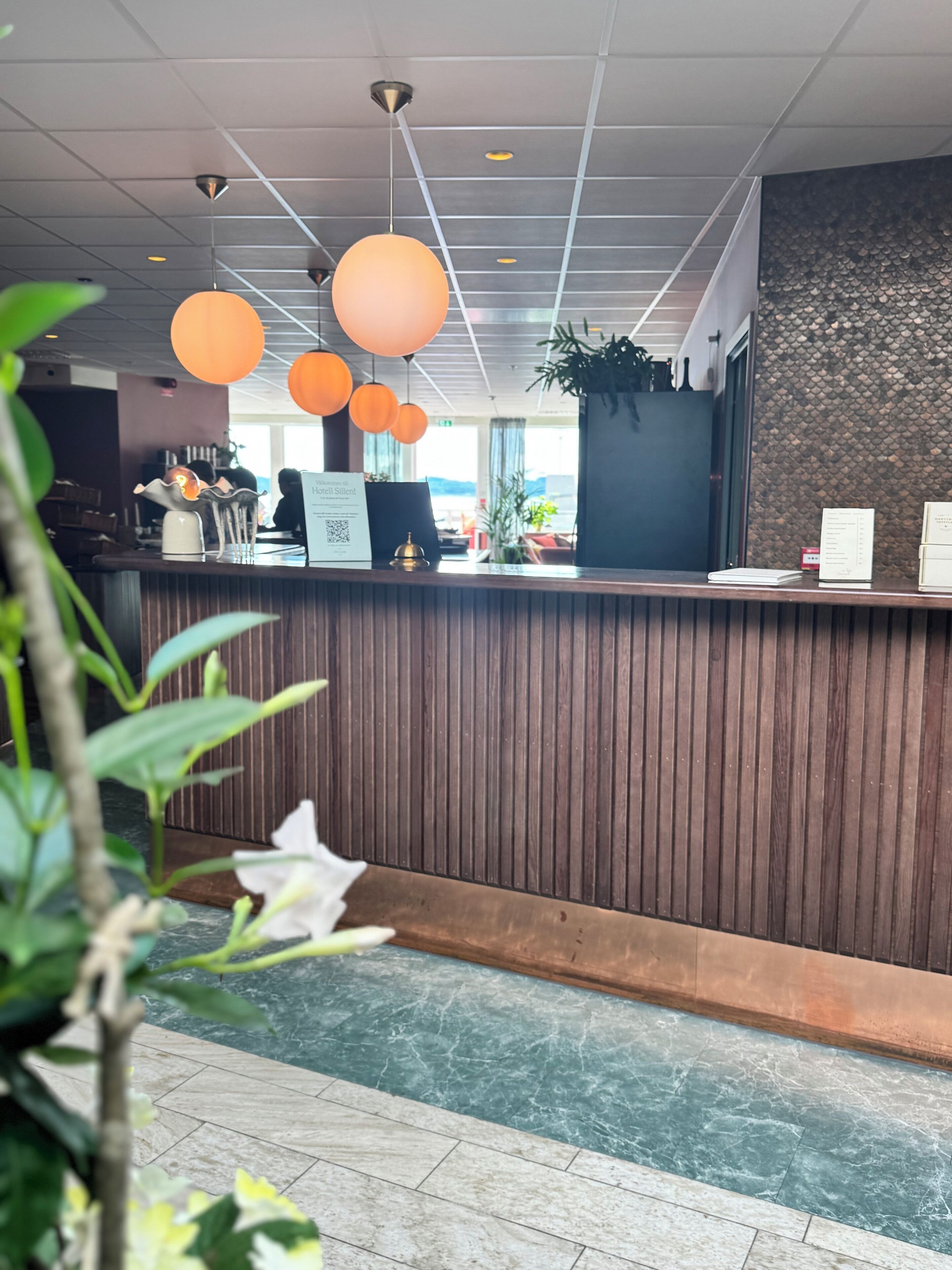Un espace d’accueil d’hôtel avec un comptoir de réception en bois, des suspensions orange, une plante en pot et une vue sur l’extérieur.