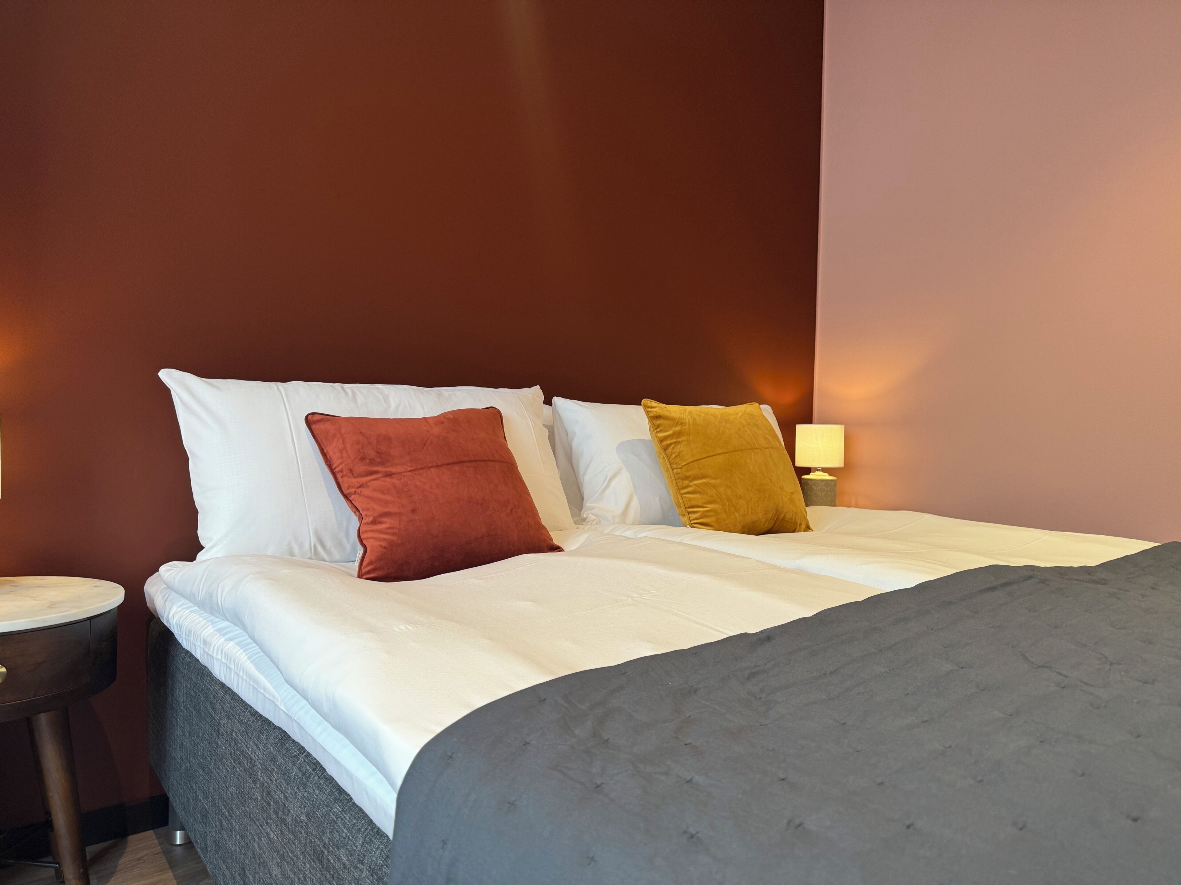 A neatly made bed with white and two pillows in shades of red and yellow, a grey blanket, and a small bedside table with a lamp.