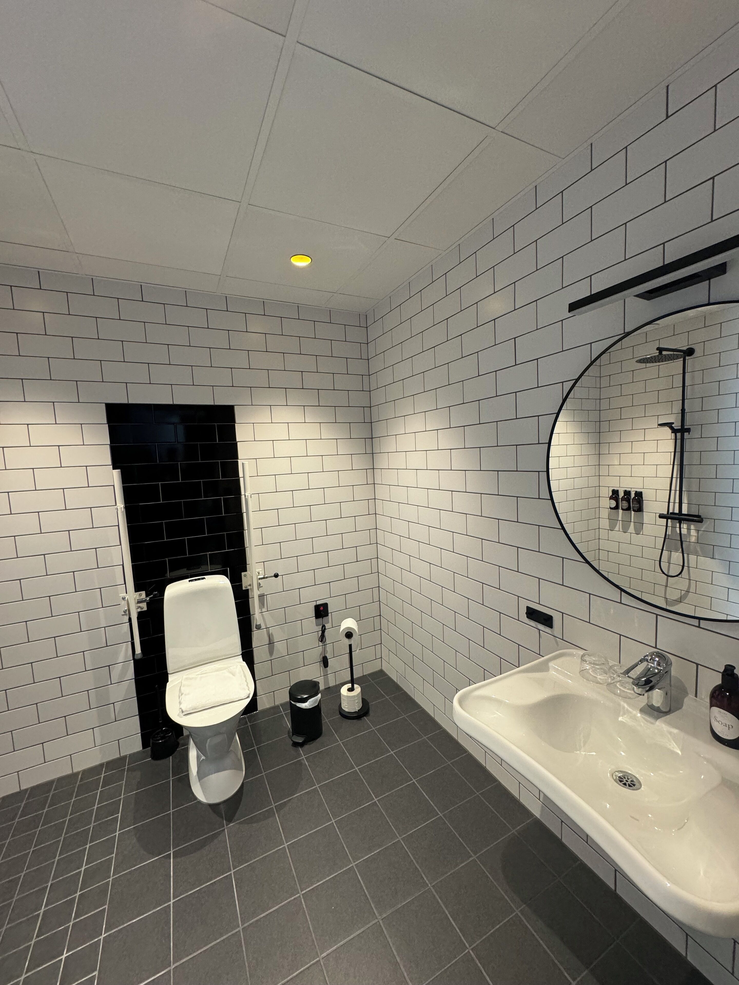 Un baño moderno con azulejos blancos tipo metro, un inodoro negro, un lavamanos blanco y una ducha con un espejo circular.