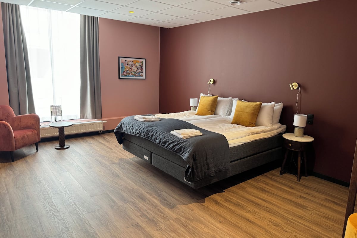 A hotel room with a large bed, a chair, a small table, and a radiator.