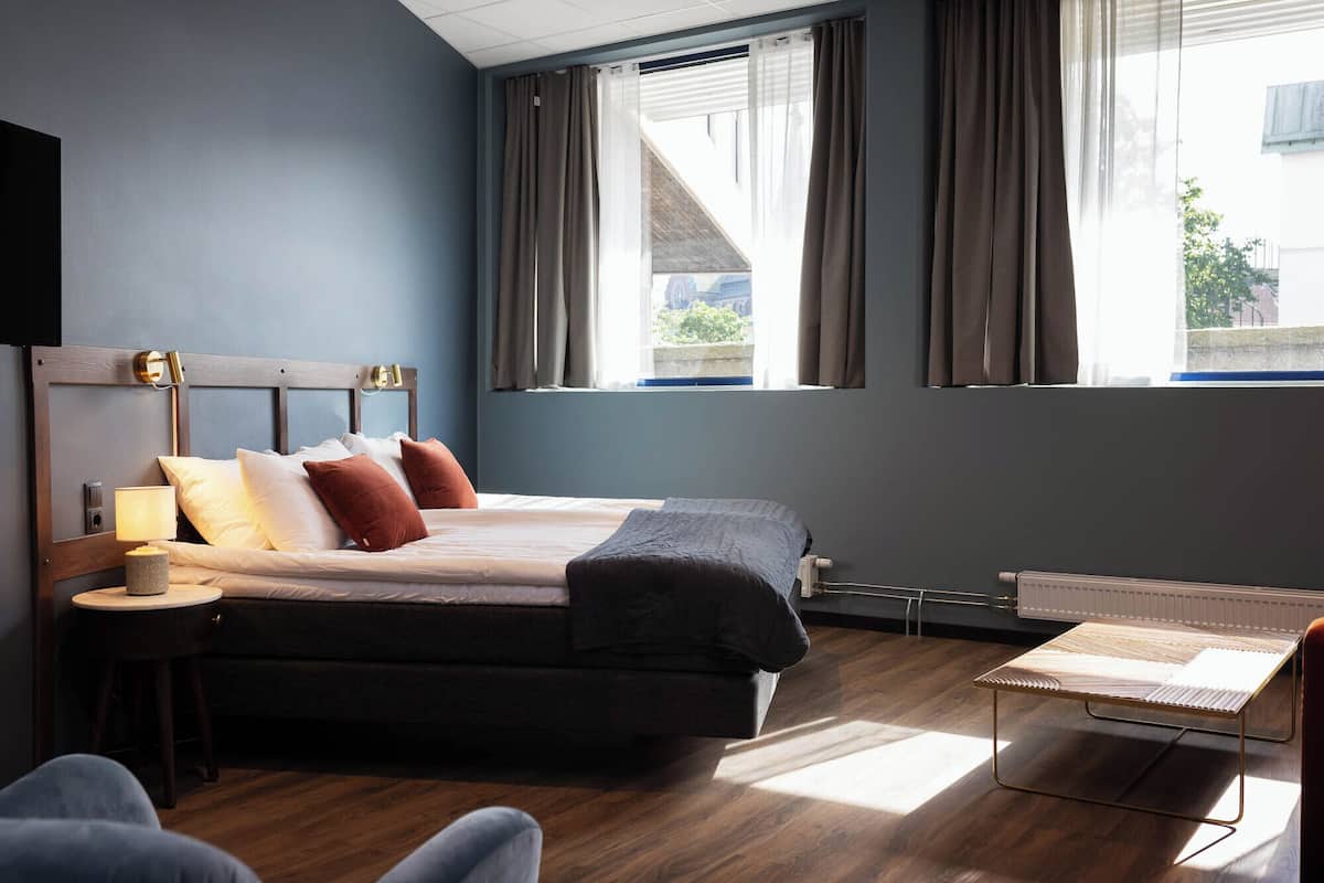 A hotel room with a bed, bedside table, lamp, and a view of the outside through large windows.