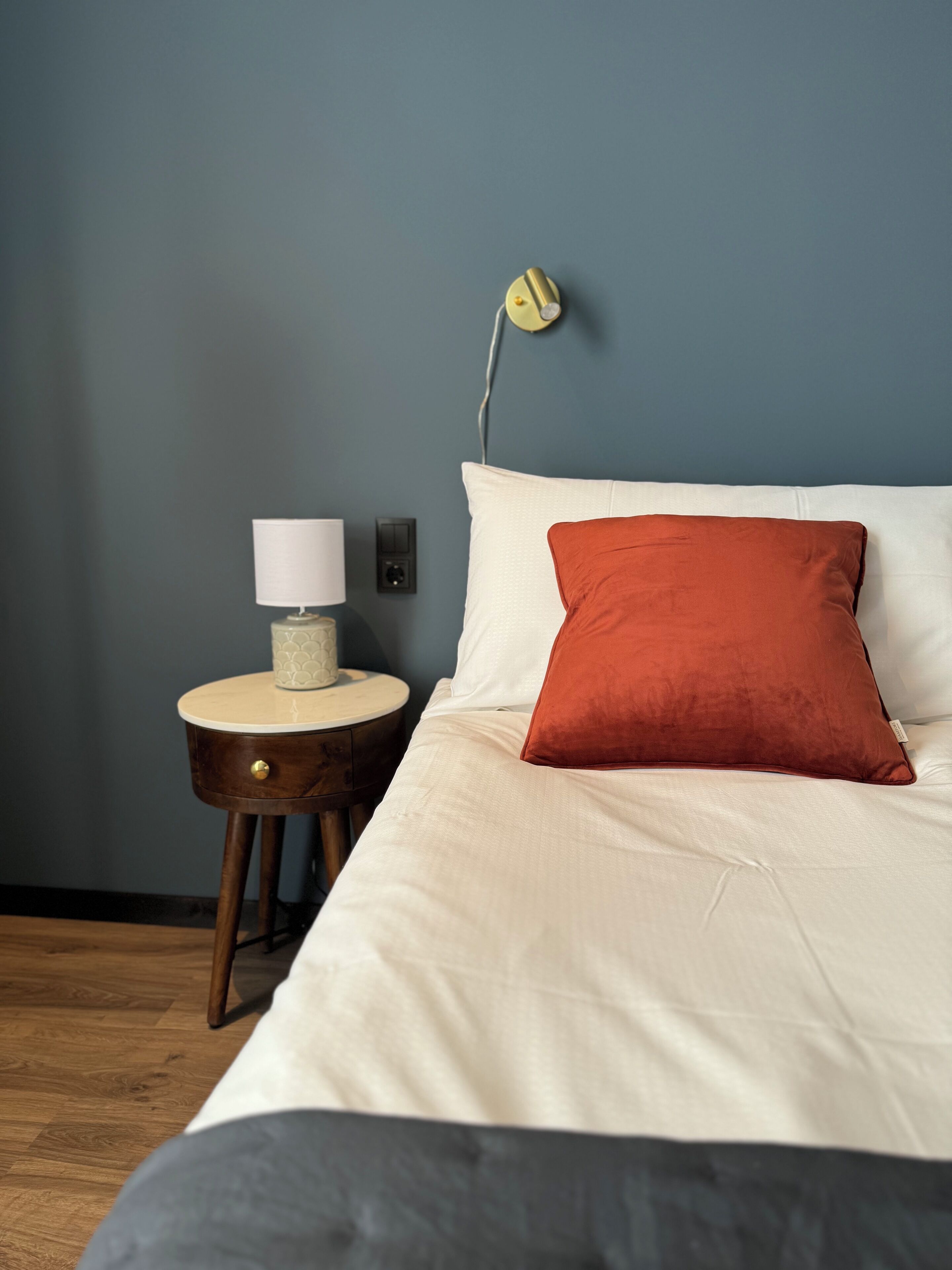 Een keurig opgemaakt bed met witte beddengoed en één rode kussen, een bedlampje, en een klein houten nachtkastje met een lamp en decoratie.