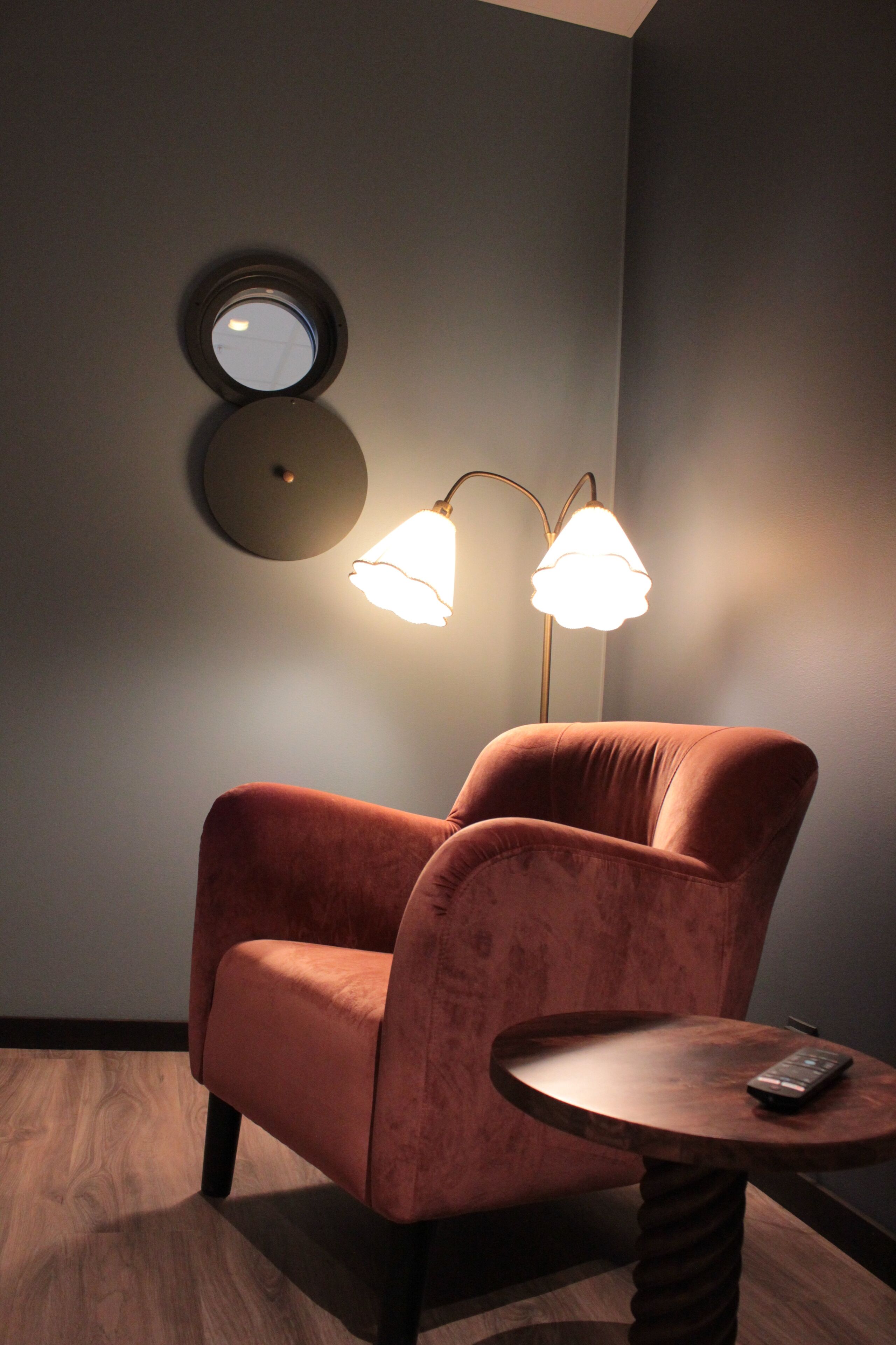A room with a velvet armchair, a small wooden table, a floor lamp, and a wall-mounted round mirror.