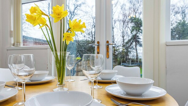 A dining table set with white plates, bowls, and wine glasses, with a vase of yellow flowers in the center. - Stunning Home Next to Nottingham University | Off-Street Parking | Sleeps 8  (Beeston)