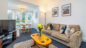 A living room with a TV, a wooden coffee table with a vase of flowers, a beige sofa with cushions, and framed pictures on the wall. - Stunning Home Next to Nottingham University | Off-Street Parking | Sleeps 8  (Beeston)