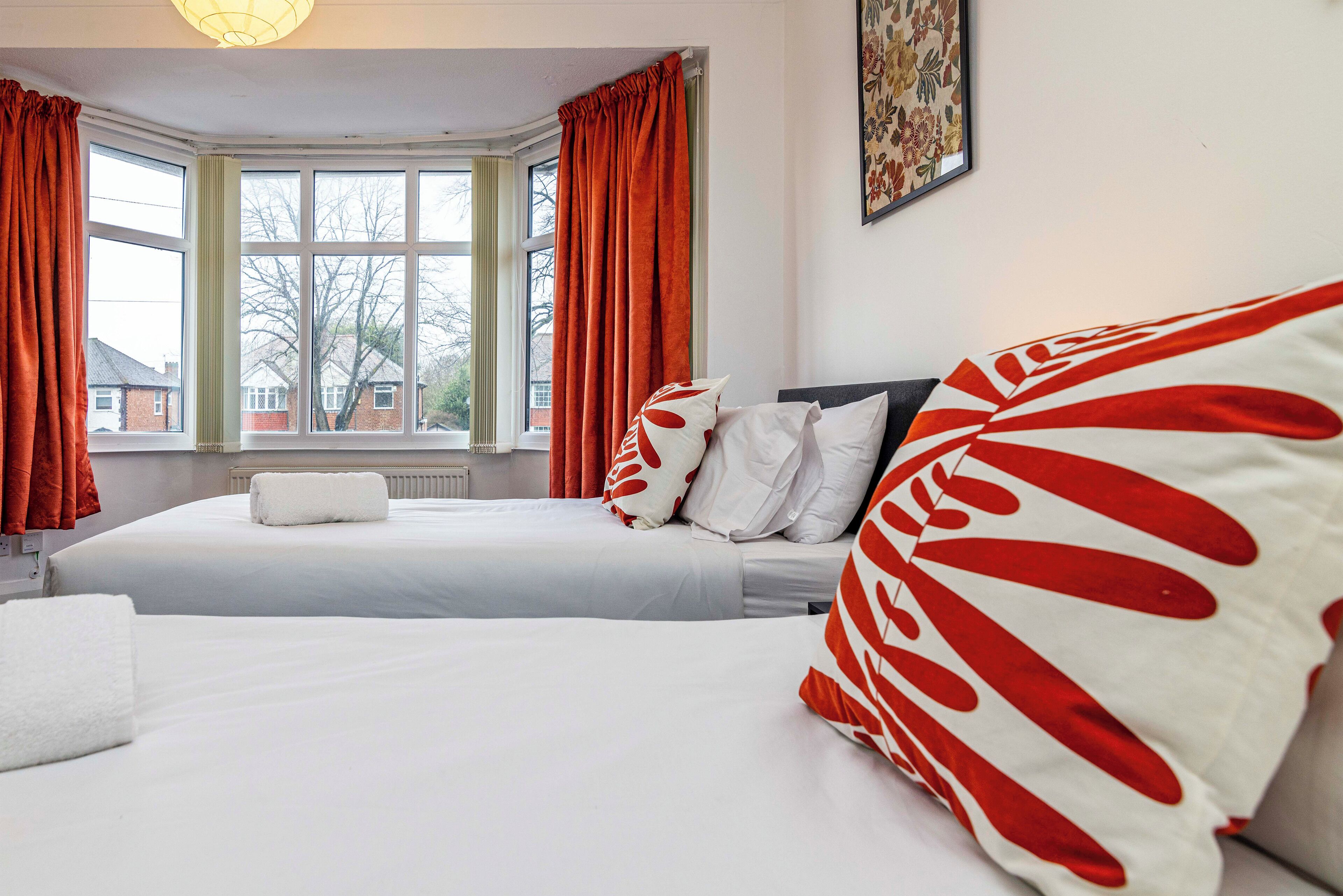 A hotel room with two beds, red curtains, white bedding, and a window with a view of houses.