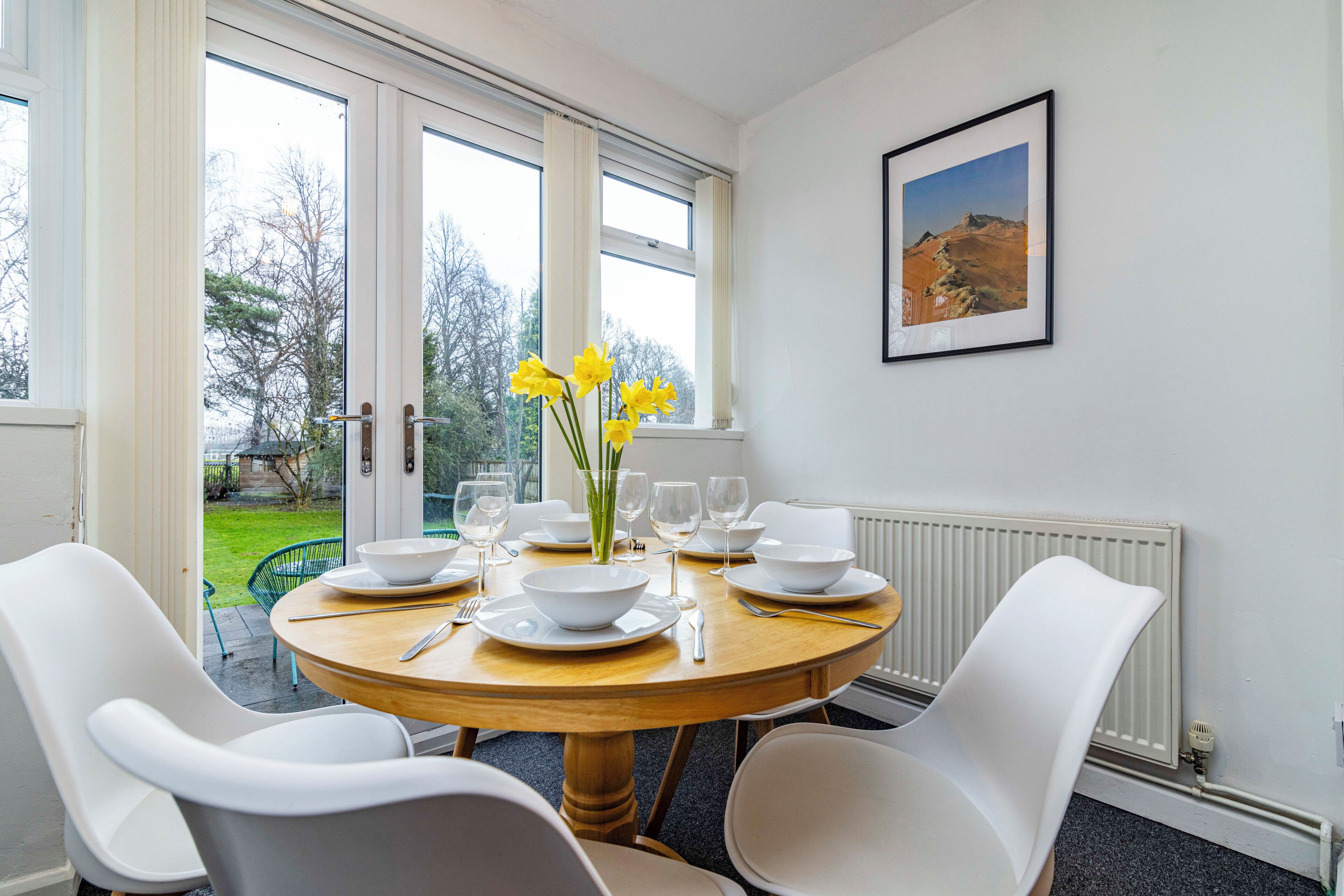 Un coin repas avec une table ronde en bois, dressée pour quatre personnes, des chaises blanches et un vase rempli de fleurs jaunes.