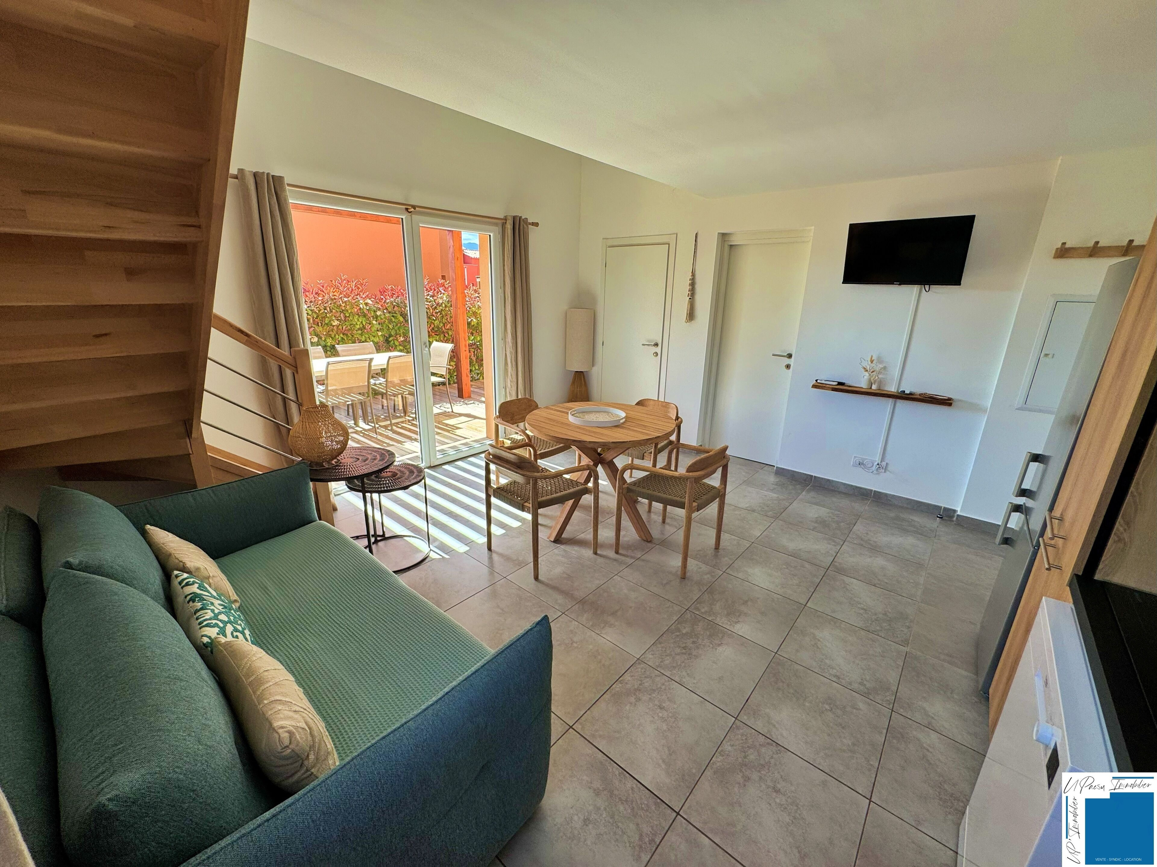 Una sala de estar moderna con un sofá, una mesa de comedor con sillas, un televisor montado en la pared y un ventanal corredero que da a un espacio exterior.