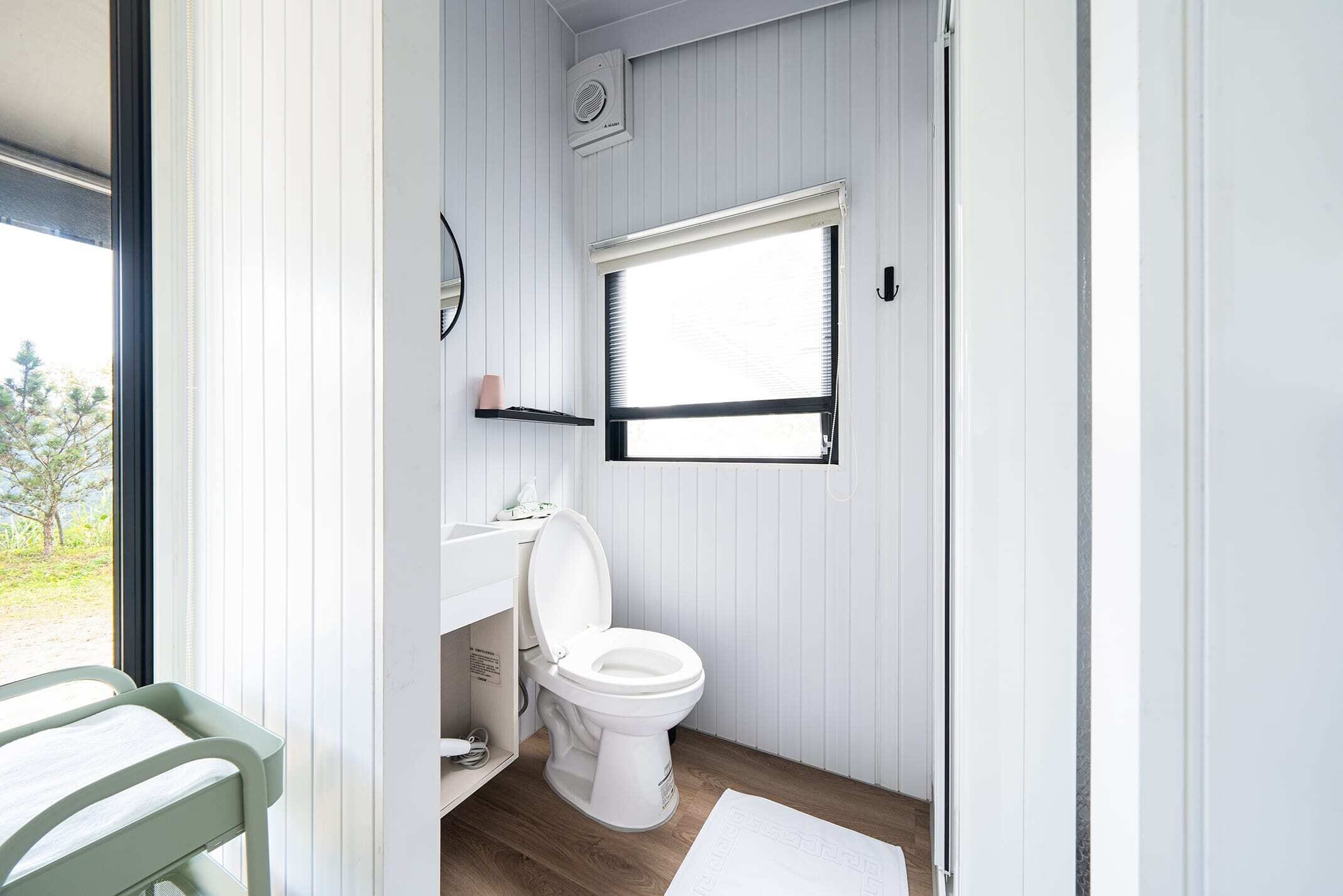 Une petite salle de bain avec des toilettes blanches, un petit lavabo et une fenêtre avec des stores.