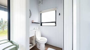 Une petite salle de bain avec des toilettes blanches, un petit lavabo et une fenêtre avec des stores.