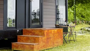 Un bâtiment moderne à l’extérieur sombre, avec un escalier en bois menant à un porche couvert et un petit espace extérieur aménagé avec une table et des chaises.
