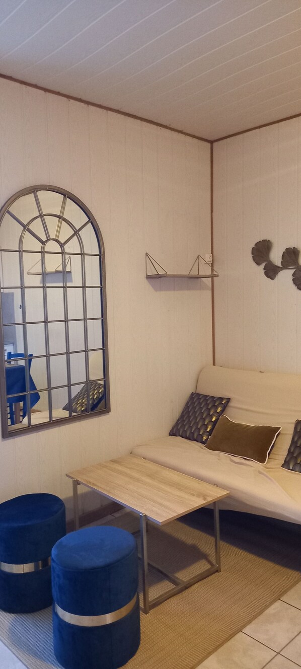 A room with a bed, a small table, two blue ottomans, a large mirror, and a decorative shelf.