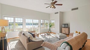 A spacious living room with a sofa, ottoman, and a TV stand. Large windows offer a view of a balcony.