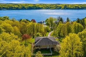 A house with a sloped roof, surrounded by lush green trees and a lake. - Private Conesus Retreat | Lake Views | Fire Pit (Conesus)