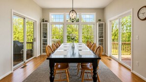 A dining room with a long table, chairs, a chandelier, and large windows with a view of greenery.