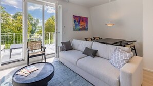 A modern living room with a sofa, coffee table, and dining table. There is a balcony with a view of trees and a painting on the wall. - Atlantic Grand Hotel - Appartement Plus mit Balkon Oder Veranda (Lübeck)