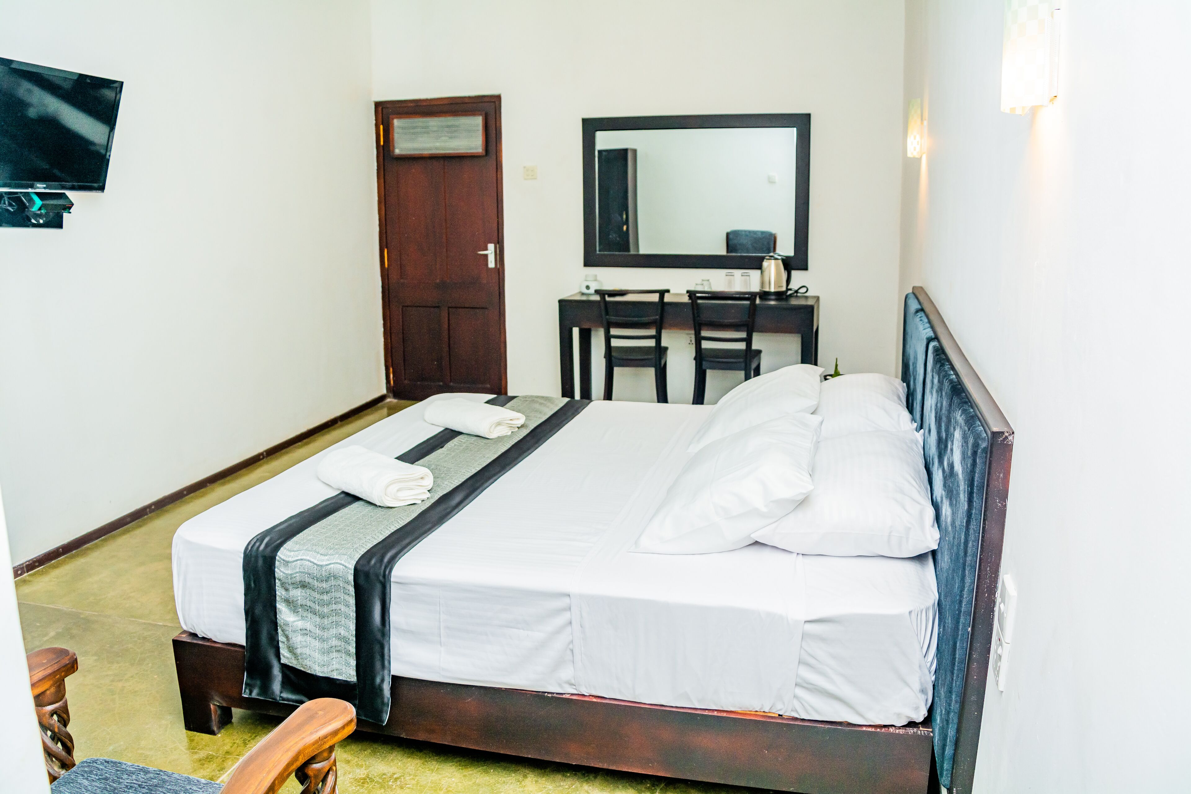 A neatly made bed with white linens and a dark headboard, a wooden desk with a chair, a flat-screen TV mounted on the wall, and a door with a wooden frame.