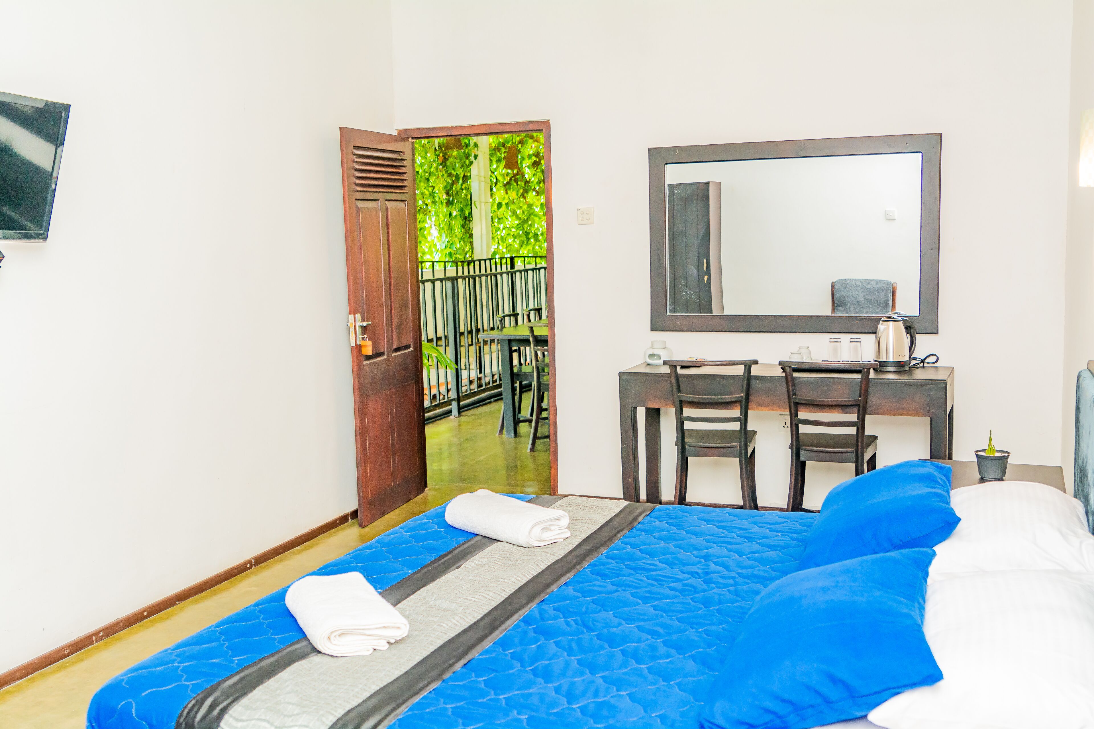 A hotel room with a bed, a desk, and a chair. There is a door leading to an outdoor area with chairs.