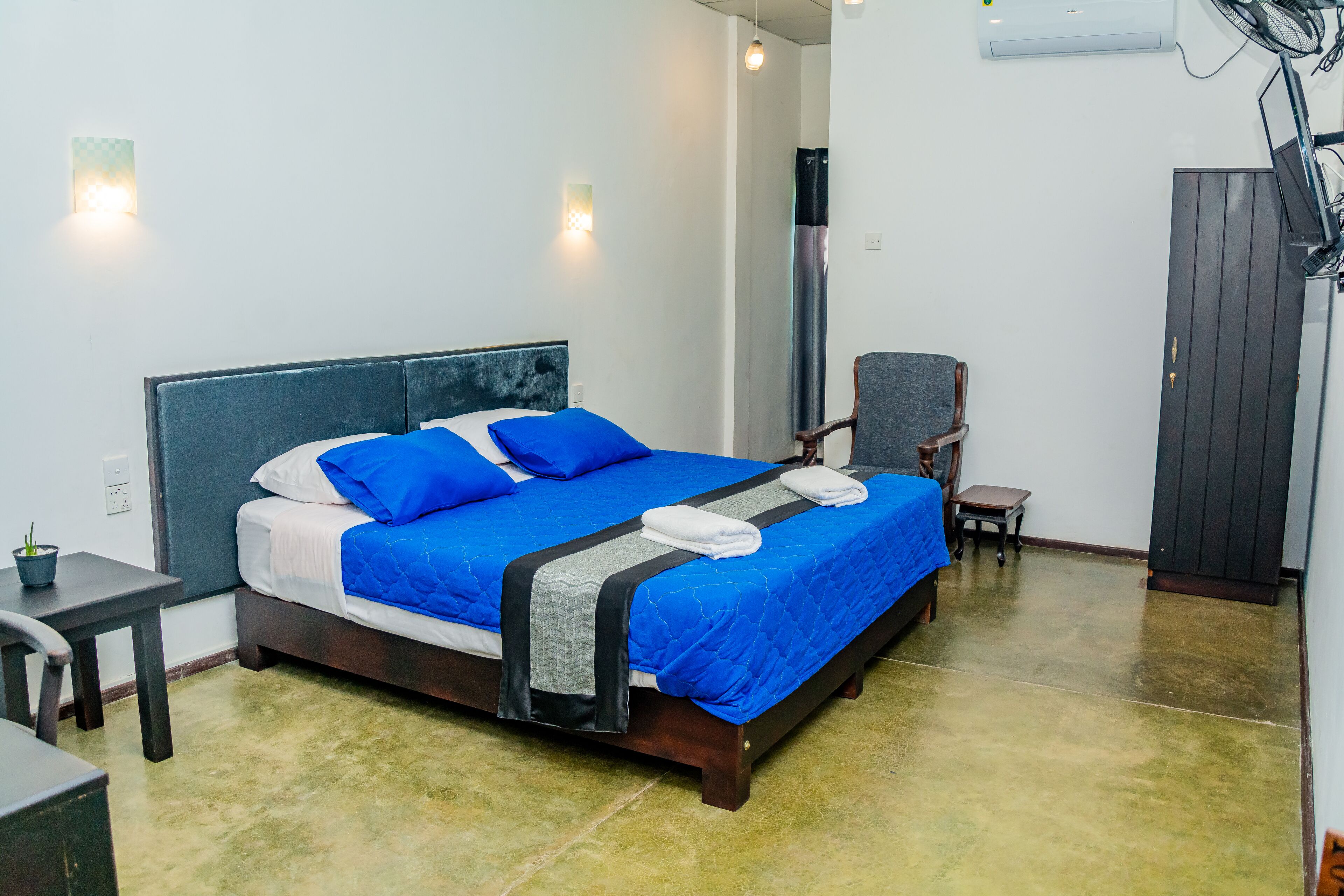 Una cama bien hecha con ropa de cama azul, una mesita con una planta y una silla en una habitación con piso de baldosas y un equipo de aire acondicionado grande en la pared.