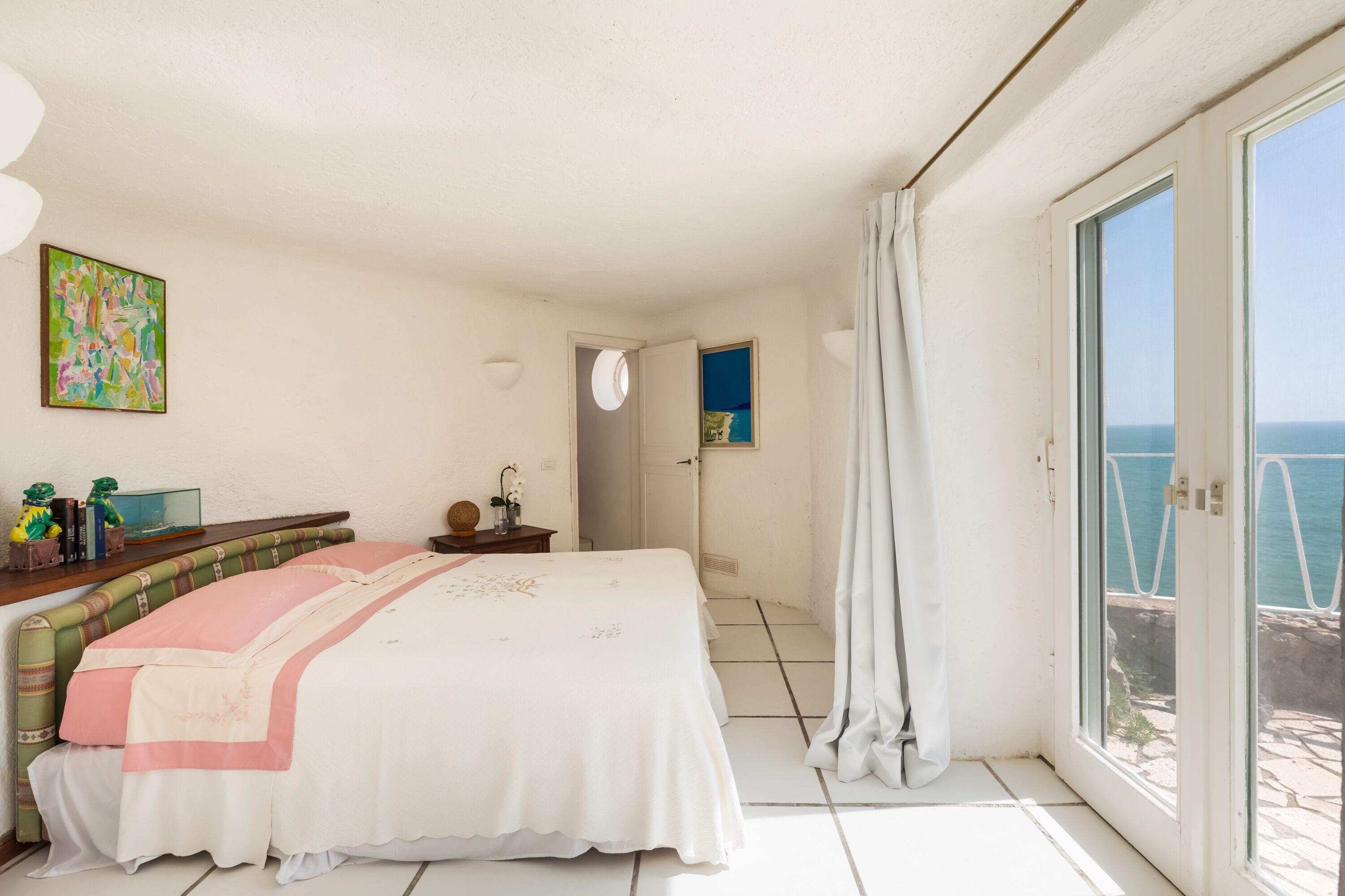 Une chambre à coucher avec un lit, une table de chevet, un tableau et une vue sur la mer par la fenêtre.