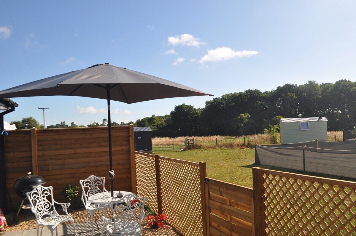 A patio with a table and chairs under a large umbrella, surrounded by wooden fencing and a view of a grassy field.