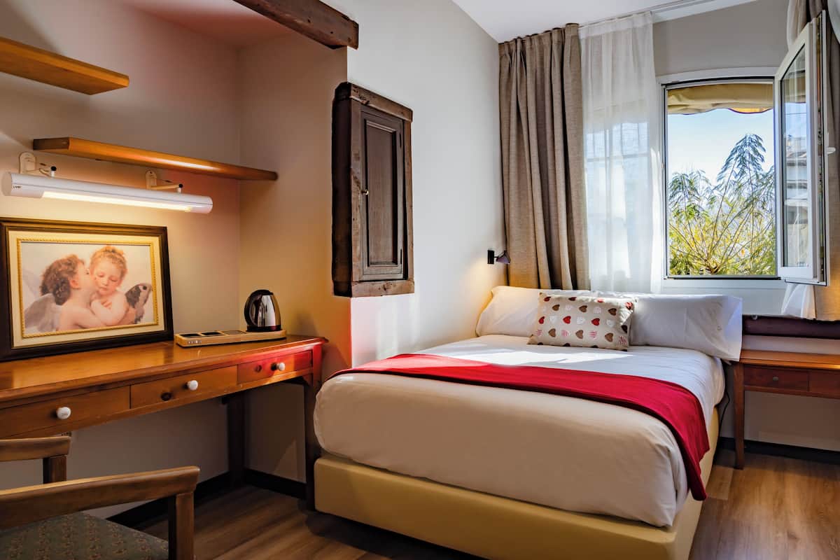 Habitación de hotel con cama, escritorio con lámpara, un marco de fotos y una ventana con cortinas.