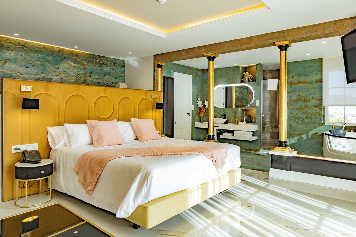 Una habitación de hotel moderna con una cama grande, una mesita de noche, un espejo y un baño visible a través de una puerta abierta.