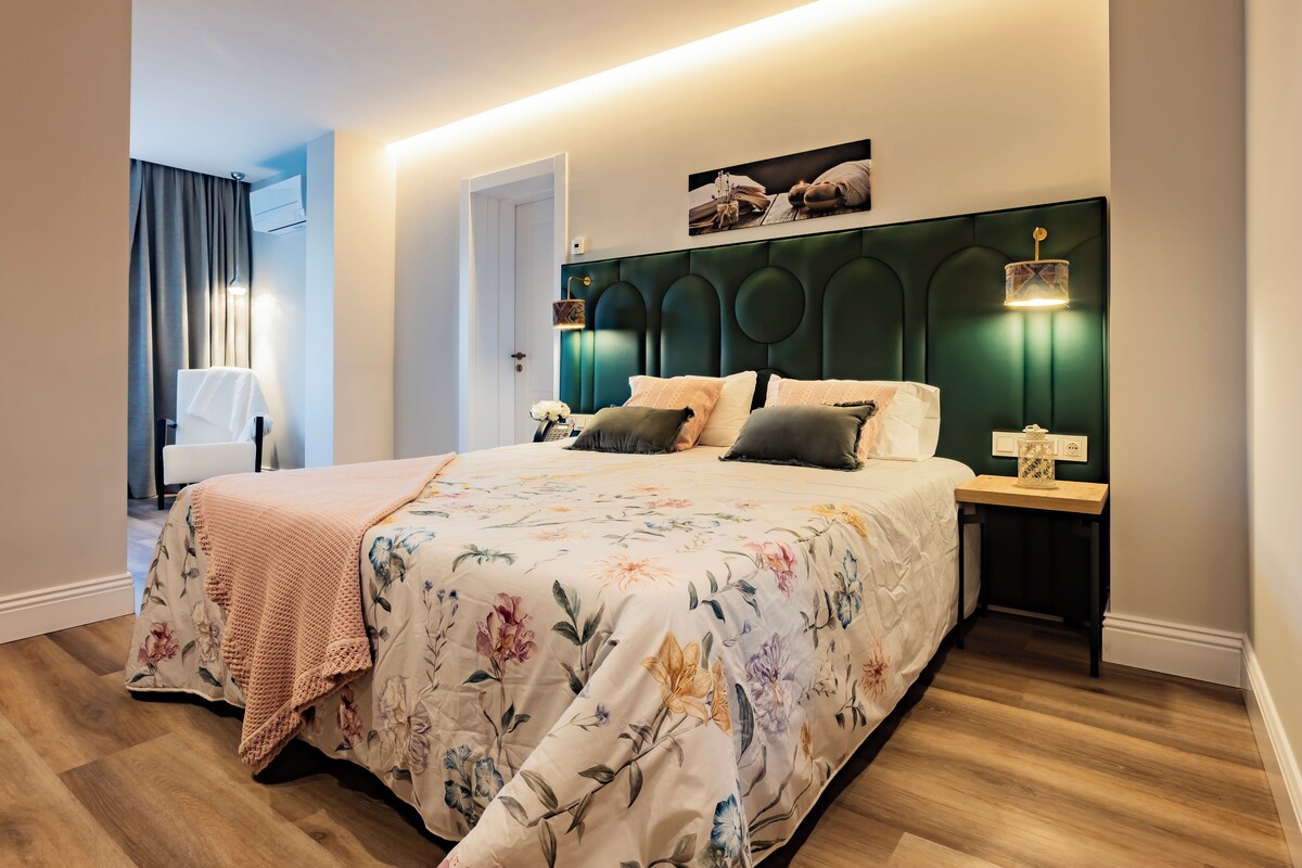 Une chambre à coucher avec une couvre-lit à motifs floraux, une tête de lit verte, des lampes de chevet et une petite table sur laquelle est posé un objet décoratif.