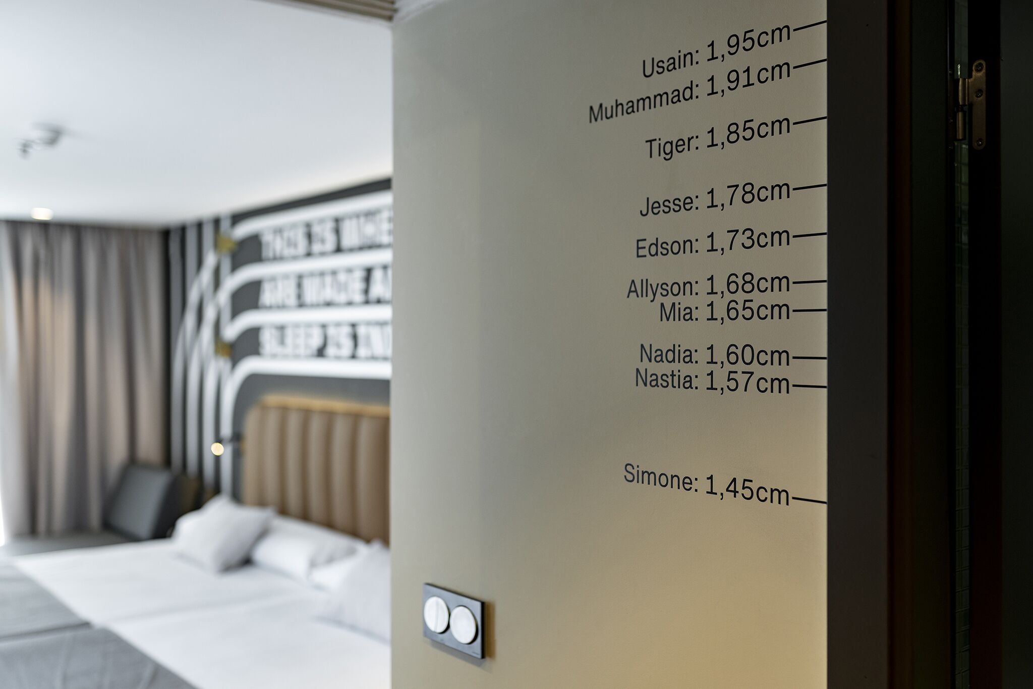 A hotel room with a bed, a wall-mounted height chart listing names and heights, and a modern design with a neutral color palette.