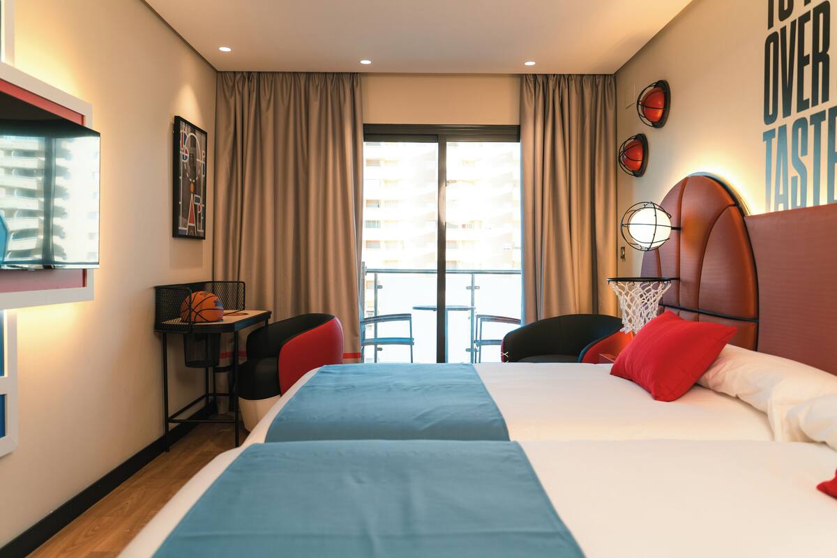 A hotel room with a bed, a desk with a basketball, a chair, a window with curtains, and a wall with a basketball hoop.