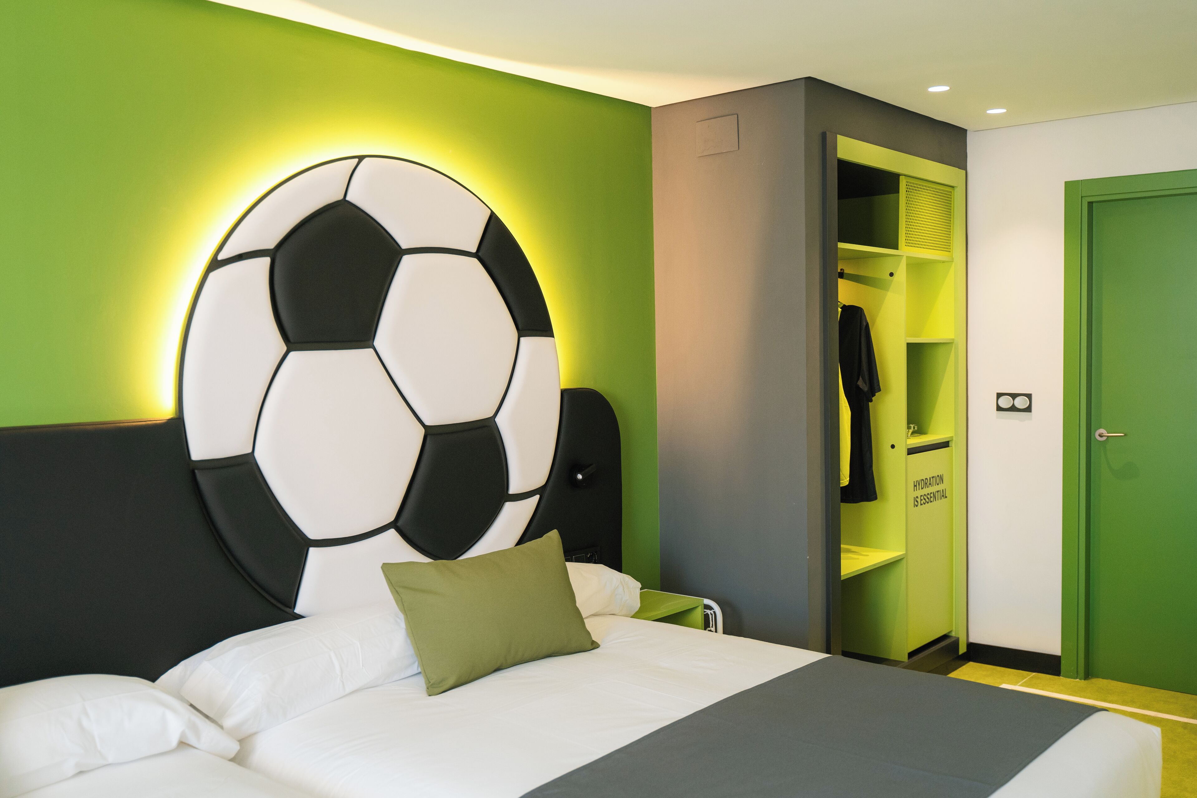 Une chambre d’hôtel avec un lit, un mur vert, une grande décoration en forme de ballon de football, une porte verte et un placard sur lequel est affiché un panneau indiquant : « L’HYDRATATION EST ESSENTIELLE ».
