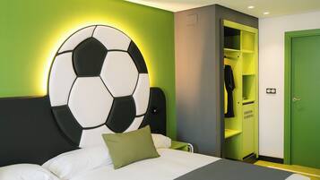 Une chambre d’hôtel avec un lit, un mur vert, une grande décoration en forme de ballon de football, une porte verte et un placard sur lequel est affiché un panneau indiquant : « L’HYDRATATION EST ESSENTIELLE ».