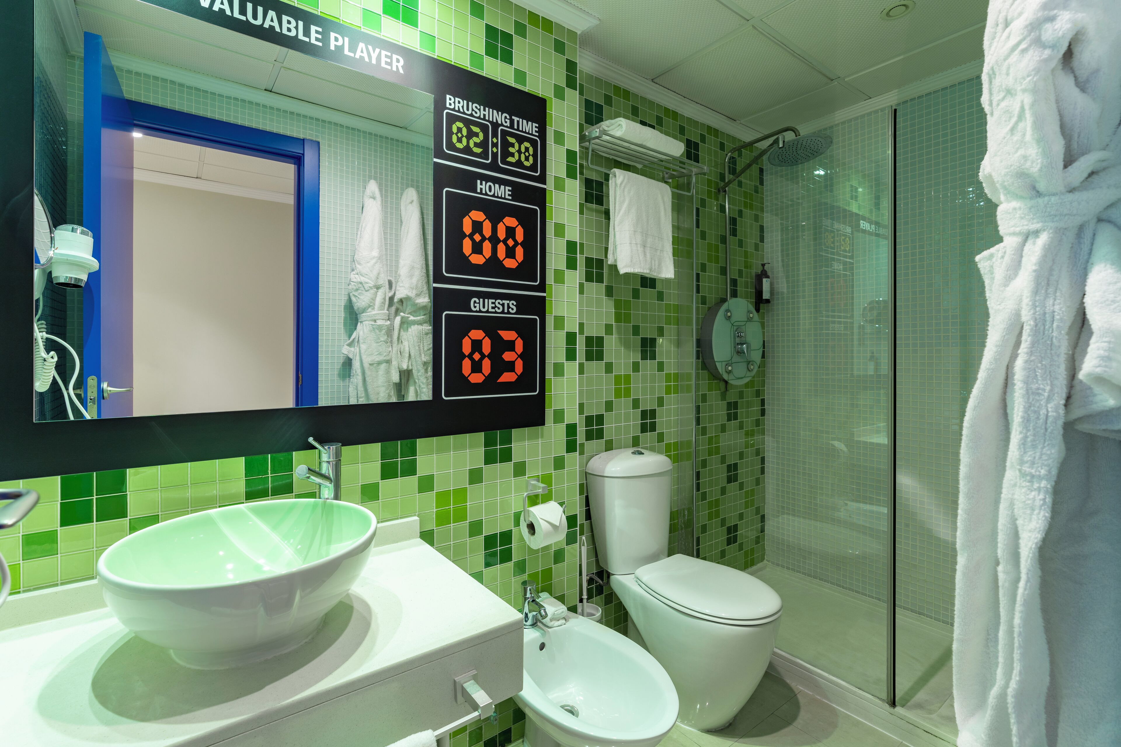 Ein Badezimmer mit grünen Fliesen, einem weißen Waschbecken, einem Bidet und einer Toilette. Oberhalb des Spiegels befindet sich eine digitale Anzeige, die Putzzeit und Bewertungen für Bewohner und Gäste zeigt.