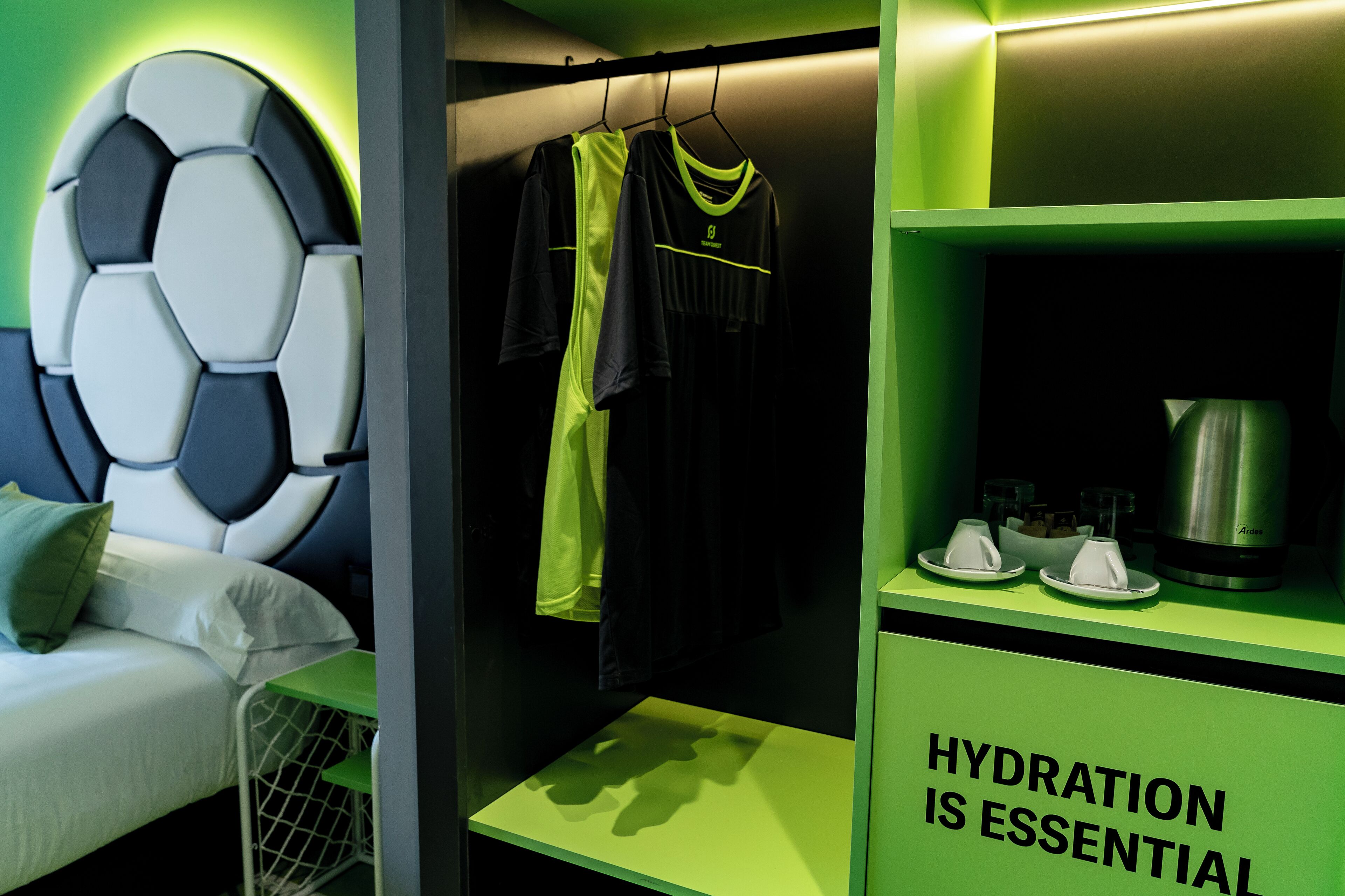 A hotel room with a bed, a wall-mounted soccer ball decoration, a rack with sports attire, a hydration sign, and a coffee machine.