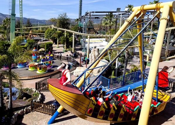 A ride with a large yellow and red boat structure, surrounded by a fenced area with palm trees and a mountainous background. - Magic Sports Hotel (Oropesa del Mar)