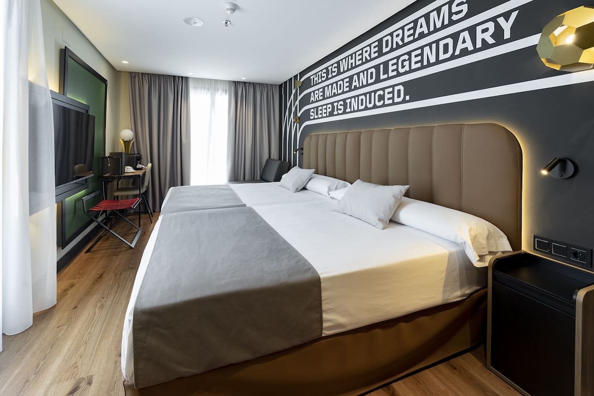 A modern hotel room with a large bed, a desk with a chair, a television, and a wall with motivational text.