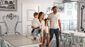 Une famille est réunie dans un coin repas moderne, avec une table et des chaises blanches, un sol à motifs et des œuvres d’art murales représentant des aliments et des ustensiles de cuisine.