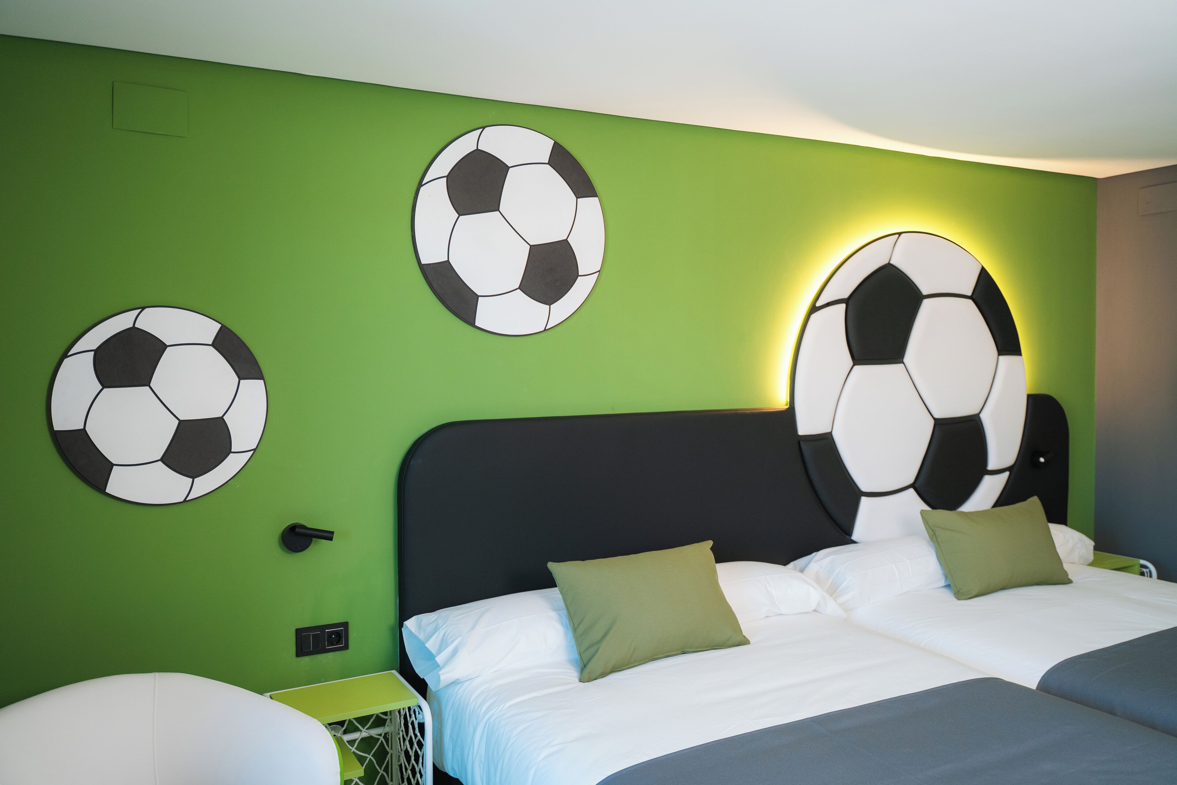 Et hotelværelse med en grøn væg, to senge med hvide sengetæpper, og tre fodboldpynt.