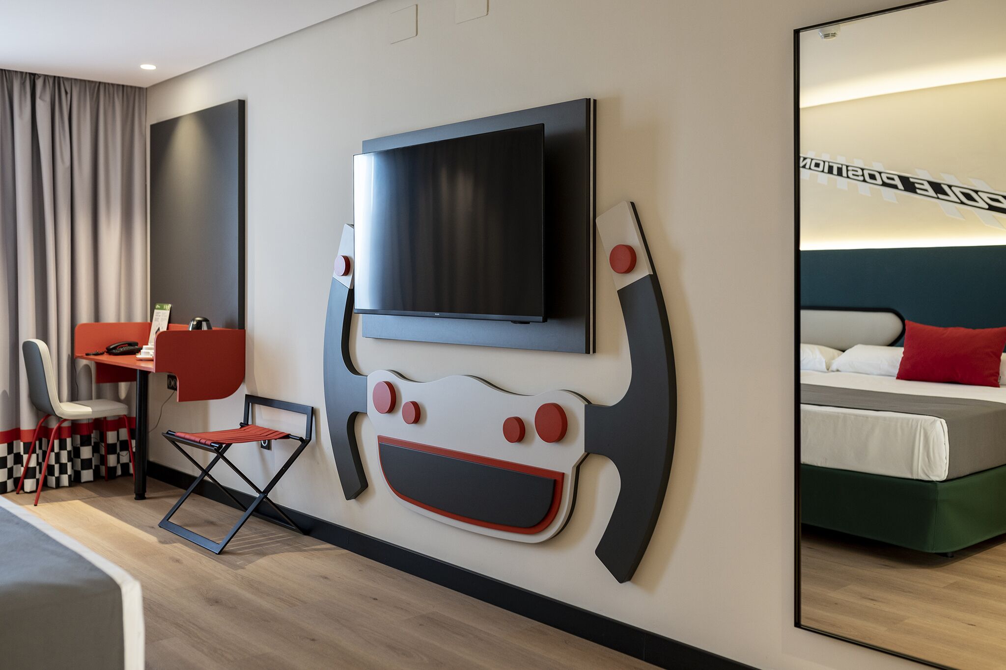 A modern hotel room with a large flat-screen TV mounted on the wall, a foldable desk, a chair, and a bed with a red pillow.