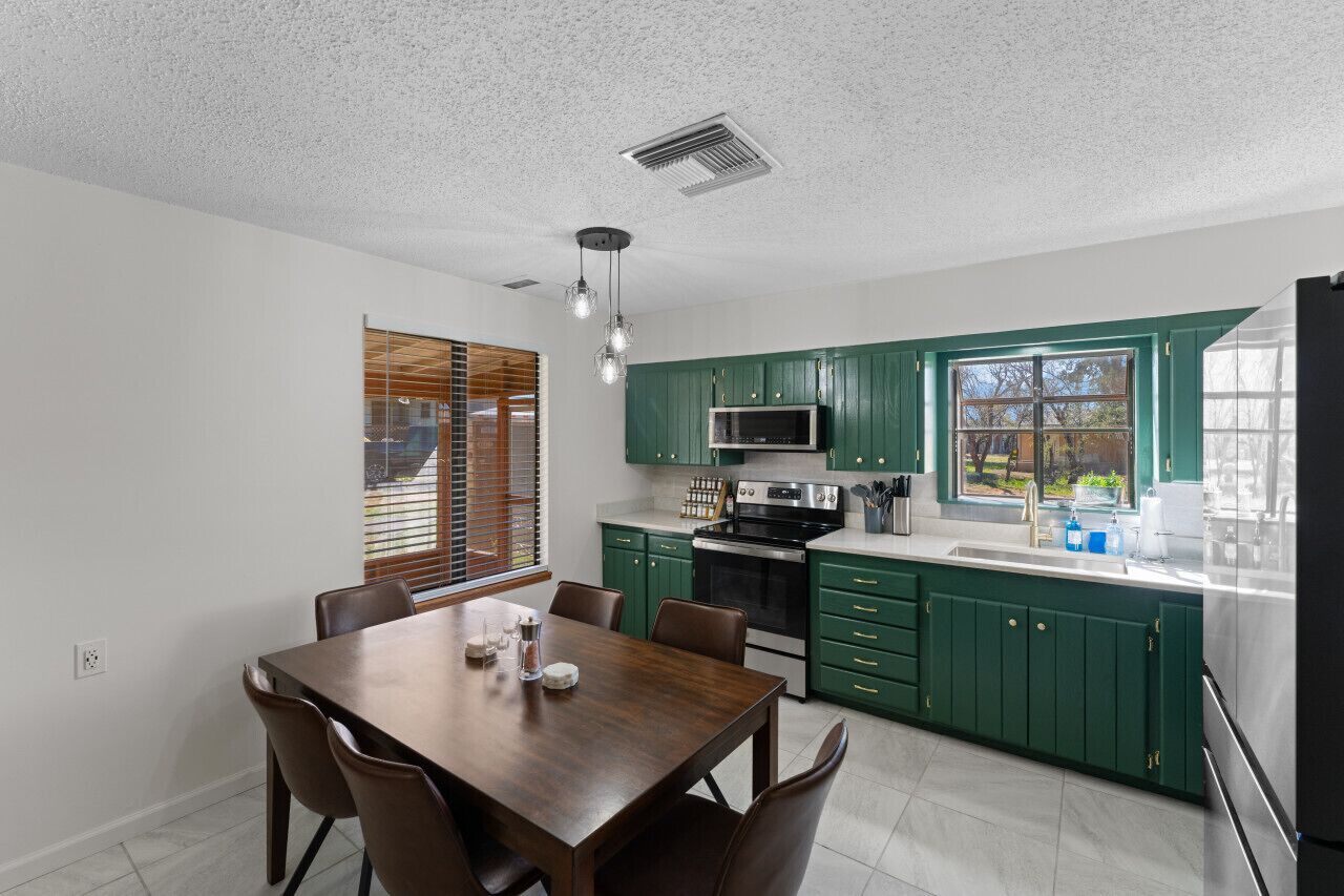 A modern kitchen with green cabinets, a dining table with chairs, and a large window with blinds.