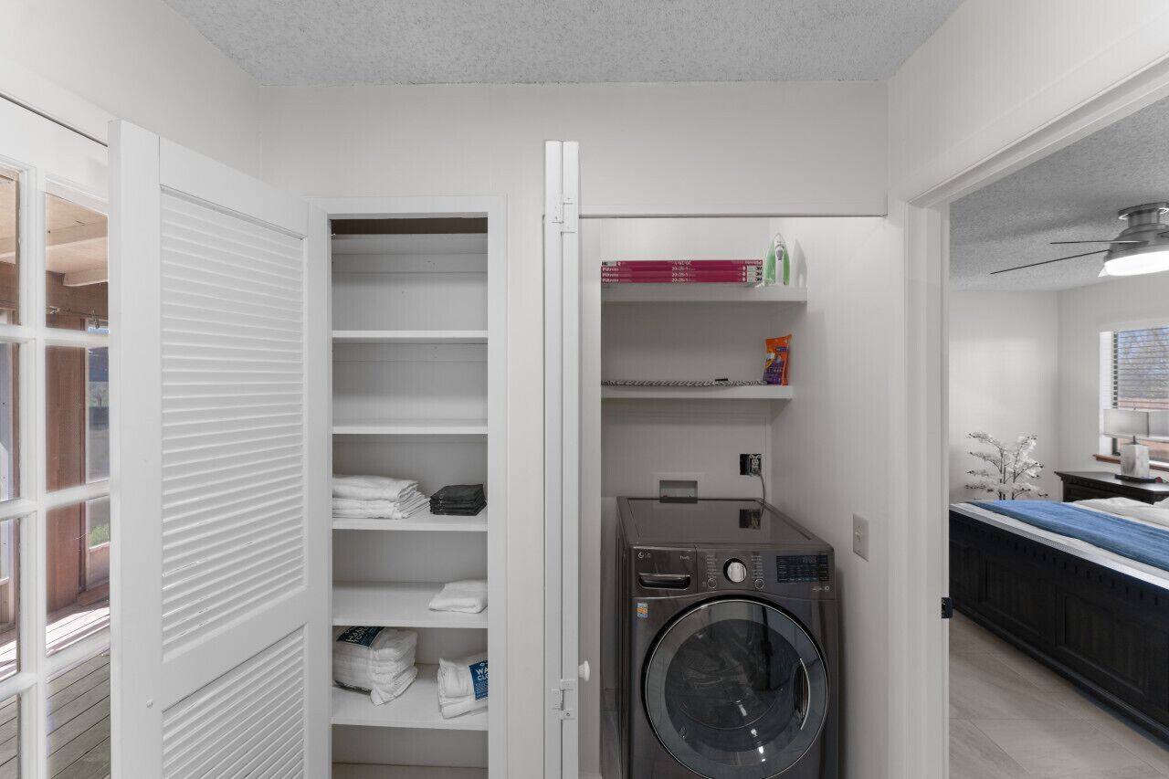 A laundry room with a washing machine, shelves with folded towels, and a window with blinds.