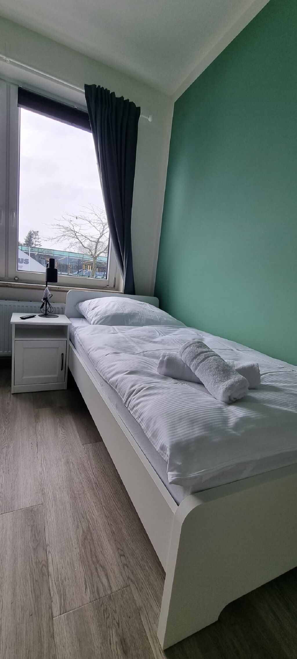 Een enkel bed met witte beddengoed, een nachtkastje met een lamp en een raam met gordijnen.