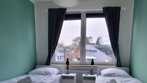 A room with two beds, a window with curtains, a hanging light fixture, and a view of buildings outside. - Zweibettzimmer in Zentraler Lage (Bremen)