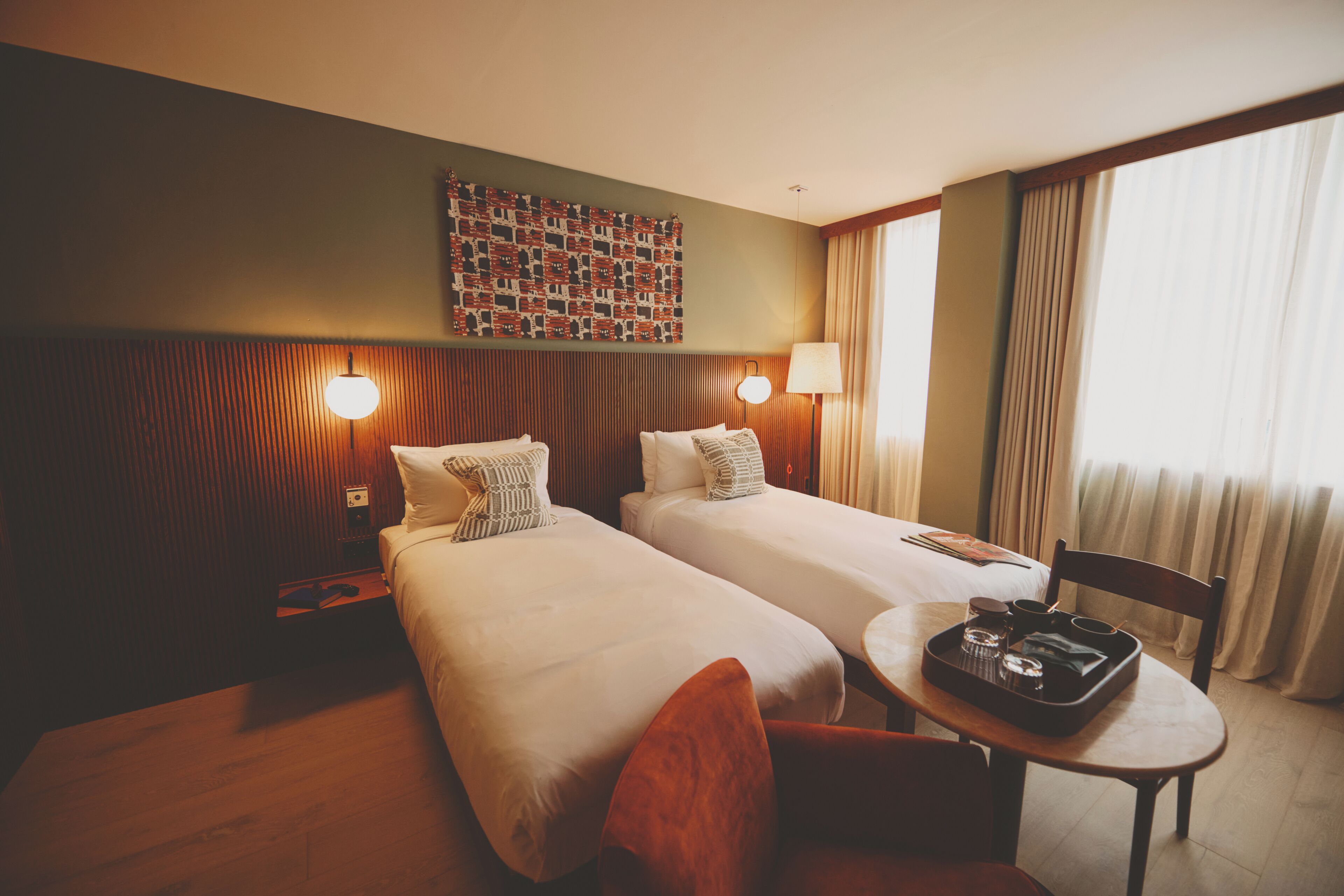 Una habitación de hotel con dos camas, una mesita con bandeja, una silla, una lámpara de pared y un adorno mural.