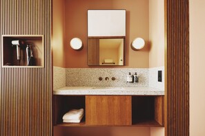 Un baño con tocador de madera, un espejo, dos luces empotradas, un perchero para toallas y un lavabo con grifo.