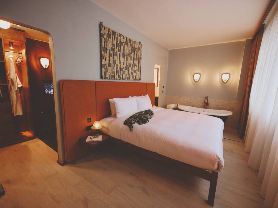 A hotel room with a bed, bedside table, lamp, artwork, and a bathroom with a sink and mirror.