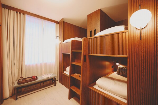 Una habitación con literas, un banco de madera, una bandeja con una taza y un plato, y una ventana con cortinas.